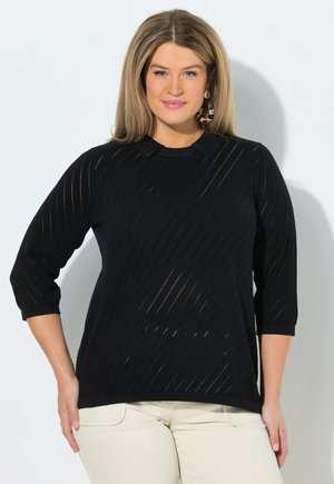Woman with straight light brown hair wears black textured sweater and beige pants, posing against a plain light background.