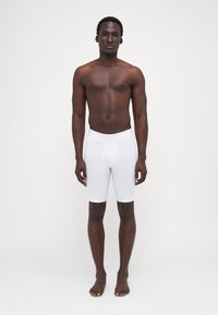 Hvide tætsiddende boxer shorts lavet af strækstof, der har en glat overflade og en bredt elastikbånd, vist af en person stående barfodet.