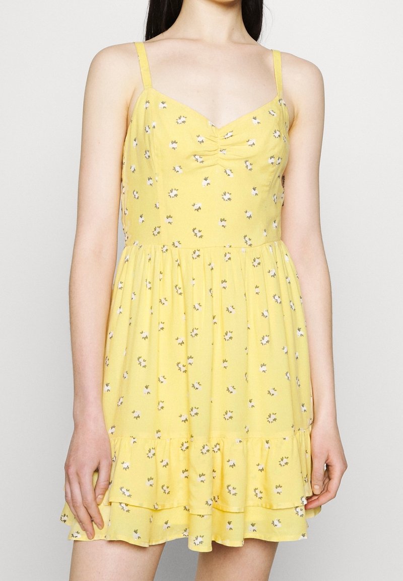 Femme portant une robe d'été sans manches jaune avec un petit motif floral blanc et un ourlet à volants.