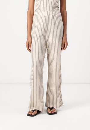 Beige wijde broek met verticale zwarte strepen, gedragen met zwarte teenslippers, getoond van taille tot voeten bij een persoon tegen een effen witte achtergrond.