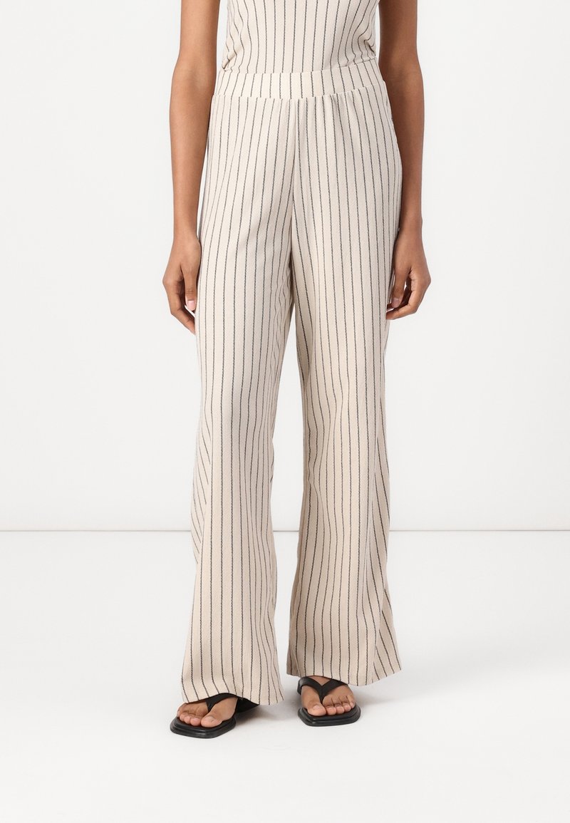 Pantalon beige à jambes larges avec des rayures noires verticales, porté avec des sandales noires, visible de la taille aux pieds sur une personne, sur un fond blanc uni.