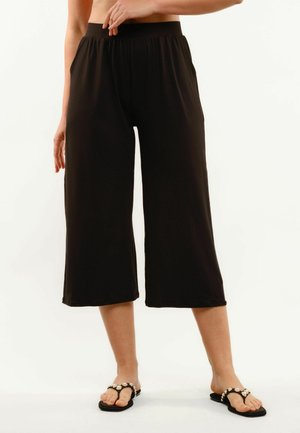 Kobieta ubrana w czarne, szerokie spodnie 7/8 z elastycznym pasem oraz czarne sandały z dużymi perłowymi ozdobami.