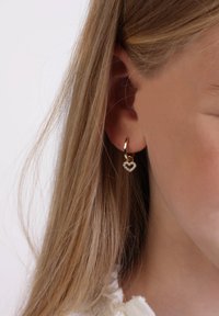 Gouden oorring met een hartvormige hanger, bezaaid met heldere edelstenen. Gekozen voor een zijprofiel met glad, steil haar.