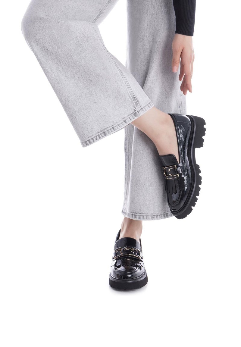 Zapatos de charol negro con suela de goma gruesa, adornados con una cadena dorada y un diseño clásico de mocasín. Se llevan con pantalones de pierna ancha grises.