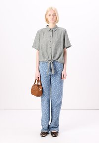 Γκρι κοντομάνικο πουκάμισο με κόμπο στη μέση, συνδυασμένο με ανοιχτό μπλε φαρδιά τζιν παντελόνια με σχέδια. Κρατάει μια μικρή καφέ τσάντα.