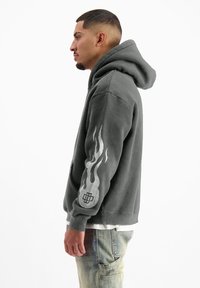 Grijze hoodie met een voorzak en een grote capuchon. Kenmerkt vlamontwerpen in zilver op de linkermouw en een logo op de manchette.