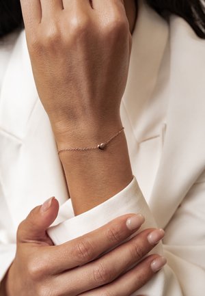 Wrist with delicate rose gold heart charm bracelet worn over white sleeve, hand gently resting on opposite arm.