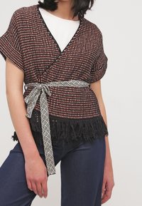 Topp i textured röd och svart mönster, med franskant och ett kontrasterande svart och vitt geometriskt bälte. Bärs med mörka jeans.