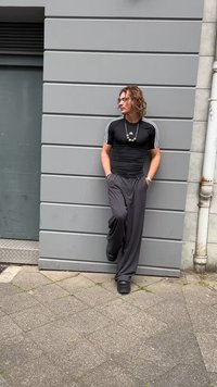 Black fitted top with white stripes on sleeves, loose gray trousers, chunky black shoes, and a beaded necklace, leaning against a gray wall.