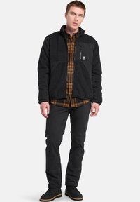 Giacca in pile nera con un pannello orizzontale e tasca con zip. Indossata sopra una camicia a quadri marrone, abbinata a pantaloni e scarpe neri.