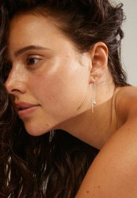 Pendientes de plata con un diseño de hilo largo y un pequeño colgante redondo. Presentan una estética elegante y minimalista. Tono de piel y textura del cabello visibles.