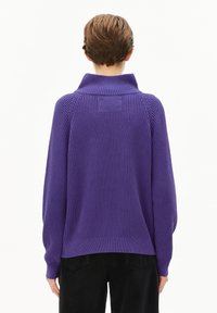 Lila gerippter Pullover mit hohem Kragen, langen Ärmeln und einem strukturierten Rückenteil, getragen über schwarzen Hosen.