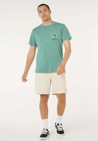 Giovane uomo che indossa una maglietta verde con stampa, pantaloncini beige, calzini bianchi e scarpe da ginnastica nere, in piedi davanti a uno sfondo semplice.