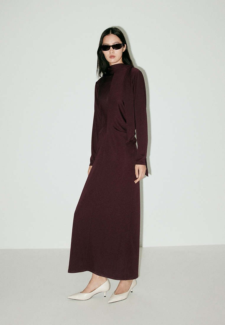 Femme portant une robe maxi manches longues violet foncé, talons blancs pointus et lunettes de soleil noires, debout devant un fond clair uni.