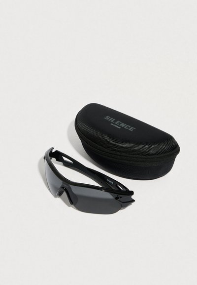 Μαύρα αθλητικά γυαλιά ηλίου με σκουρόχρωμους φακούς τοποθετημένα δίπλα σε μαύρη σκληρή θήκη που φέρει την ετικέτα «SILENCE» σε λευκό φόντο.