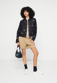 Tmavomodrá rifľová bunda s vyšívanými vzormi, béžový top, khaki šortky, biele tenisky a modrá kabelka. Model pózuje na neutrálnom pozadí.