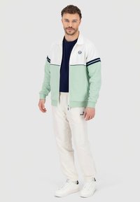 Ljusgrön zip-up jacka med vita och marinblå detaljer, bärs över en marinblå tröja, kombinerad med crèmefärgade mjukisbyxor och vita sneakers.