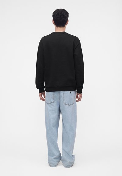 Homme de dos portant un sweat-shirt noir à manches longues et un jean ample bleu clair sur un fond blanc uni.