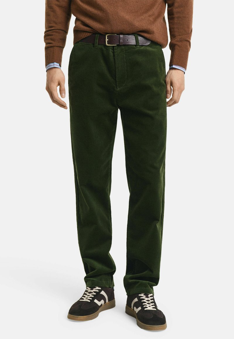 Grüne Cordhosen mit geradem Bein, ausgestattet mit einem vorderen Knopfverschluss und seitlichen Taschen. Getragen mit einem braunen Pullover und Sneakers.