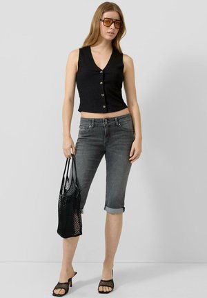 Femme portant un haut noir sans manches boutonné, un jean capri gris retroussé, des talons noirs en résille, des lunettes de soleil, tenant un sac cabas noir en résille.