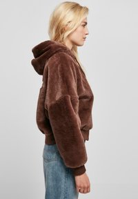 Haut à capuche en fausse fourrure marron avec une coupe décontractée, des poignets côtelés et une capuche, présentant une texture douce et un ourlet marron contrastant.