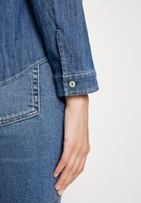 Close-up de uma jaqueta de denim, mostrando o tecido azul texturizado, mangas dobradas com costura leve e um botão verde em destaque perto da manga.