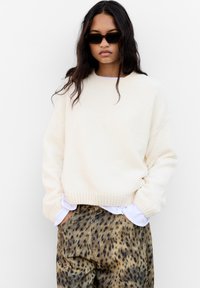 Pull en tricot côtelé crème avec une coupe décontractée, associé à un pantalon marron à imprimé léopard. Le mannequin porte des lunettes de soleil noires.