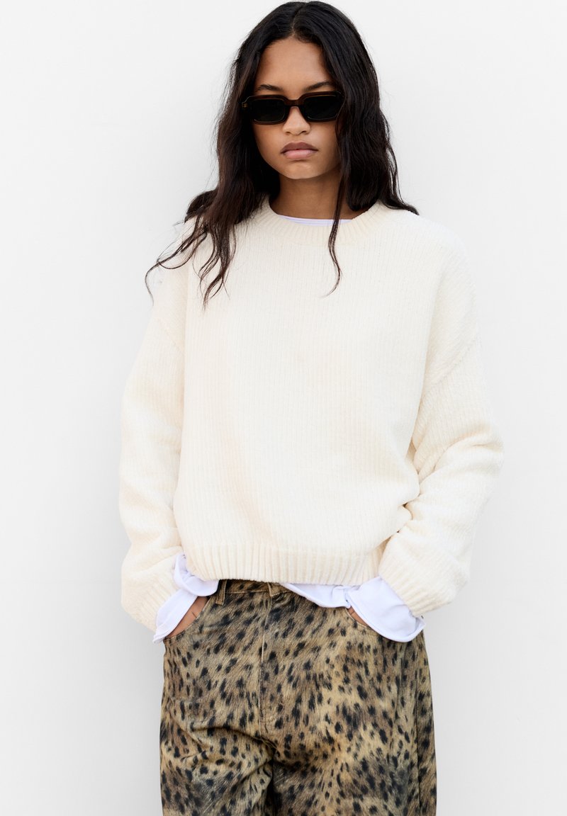 Pull en tricot côtelé crème avec une coupe décontractée, associé à un pantalon marron à imprimé léopard. Le mannequin porte des lunettes de soleil noires.