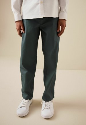 Person wearing dark green straight-leg pants with white sneakers and a white button-up shirt, standing on a beige floor.