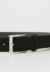 Curea din suede negru cu cataramă din metal argintiu. Prezintă o textură netedă și mai multe găuri de ajustare. Logo-ul este embossat pe cataramă.