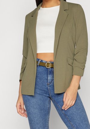 Kobieta w oliwkowej marynarce na białym krótkim topie, do tego niebieskie dżinsy z wysokim stanem i oliwkowy pasek ze złotą klamrą.