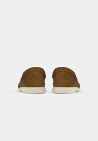 Zapatos marrones de ante sin cordones con punta redonda, detalles de costura sutiles y suela de goma blanca. Diseño minimalista con un perfil bajo.
