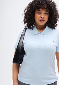 Ανοιχτό μπλε πόλο με κλασικό γιακά, πέντε κουμπιά και μικρό λογότυπο. Φοριέται με μαύρη τσάντα ώμου. Λιτή υφή.