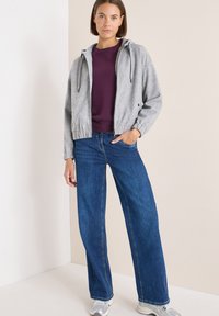 Chaqueta gris con cremallera y capucha, suéter morado debajo y pantalones vaqueros de pierna ancha azul; textura suave, diseño casual y paleta de colores neutra.
