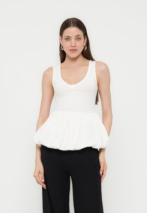 Femme aux longs cheveux bruns portant des boucles d’oreilles créoles dorées, un haut blanc sans manches avec un ourlet péplum, et un pantalon noir, debout devant un fond blanc.