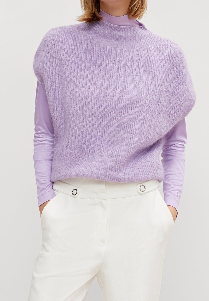 Femme portant un col roulé lavande et un pull sans manches en tricot lilas doux et texturé sur un pantalon blanc taille haute, les mains dans les poches.