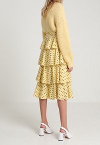 Donna che indossa un maglione spesso giallo e una gonna a strati gialla con pois neri, abbinata a scarpe con tacco a gamba bianca.