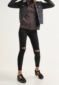 Woman wearing black shearling-collar jacket, maroon sweater, ripped black skinny jeans, and black platform Mary Jane shoes.