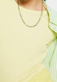 Haut côtelé jaune avec un collier en chaîne argentée, superposé sous une chemise rayée verte clair et blanche. Texture douce, design ajusté.