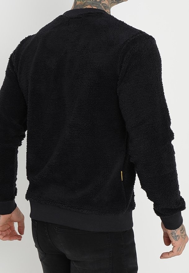 Homme portant un pull noir duveteux à manches longues, vu de dos, avec des tatouages visibles sur le cou et les mains.