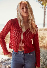 Cardigan lavorato a maglia rosso con un motivo a zigzag, lunghezza cropped, maniche lunghe e chiusura con nastro, abbinato a jeans blu a vita alta.
