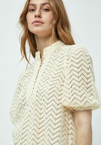 Krämfärgad blus med ett zigzag-mönster, korta puffärmar och en hög halsringning med knappslutning fram.