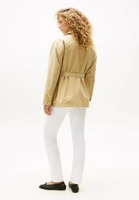 Beige lichtgewicht jack met een ceintuur in de taille, lange mouwen en knoopaccenten, gecombineerd met witte aansluitende broek en zwarte platte schoenen.