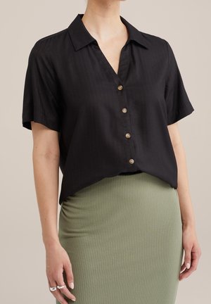 Vrouw draagt een zwart katoenen blouse met korte mouwen en knopen, met een kraag, en een geribde olijfgroene rok, met ringen aan de linkerhand.