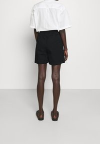 Person standing facing away wearing black shorts, white short-sleeve shirt, and black slip-on shoes against plain background.