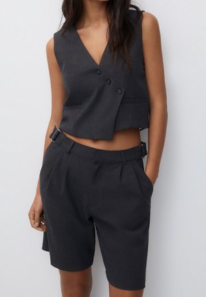Femme portant un gilet court sans manches, asymétrique gris foncé et un short taille haute assorti avec des détails de ceinture.