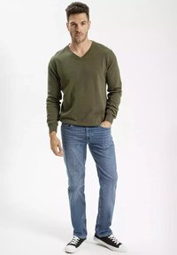 Zielona oliwkowa sweter z dekoltem w serek, wykonany z dzianiny, w połączeniu z jasnoniebieskimi dżinsami. Czarno-białe sneakersy z teksturowanymi podeszwami.