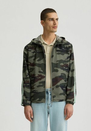 Veste zippée camouflage dans des nuances de vert variées avec une capuche, des poignets élastiques et des accents de rayures blanches sur les manches, associée à un jean bleu clair.