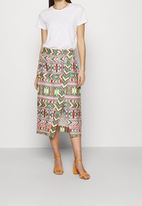 Gonna multicolore con motivo a fantasia e design avvolgente, caratterizzata da forme geometriche in rosa, verde e rosso. Abbinata a una t-shirt bianca semplice e tacchi gialli.