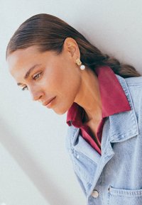 Guld- och pärlörhängen med rektangulär design. Bärs av en kvinna med långt brunt hår, kompletterad med en denimjacka och en burgunderfärgad skjorta.